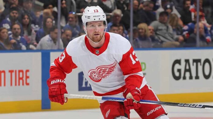 Detroit Red Wings forward Alexis Lafrenière, wearing number 13 in a white away jersey with red trim and the winged wheel logo, skates on the ice holding his stick during a game.