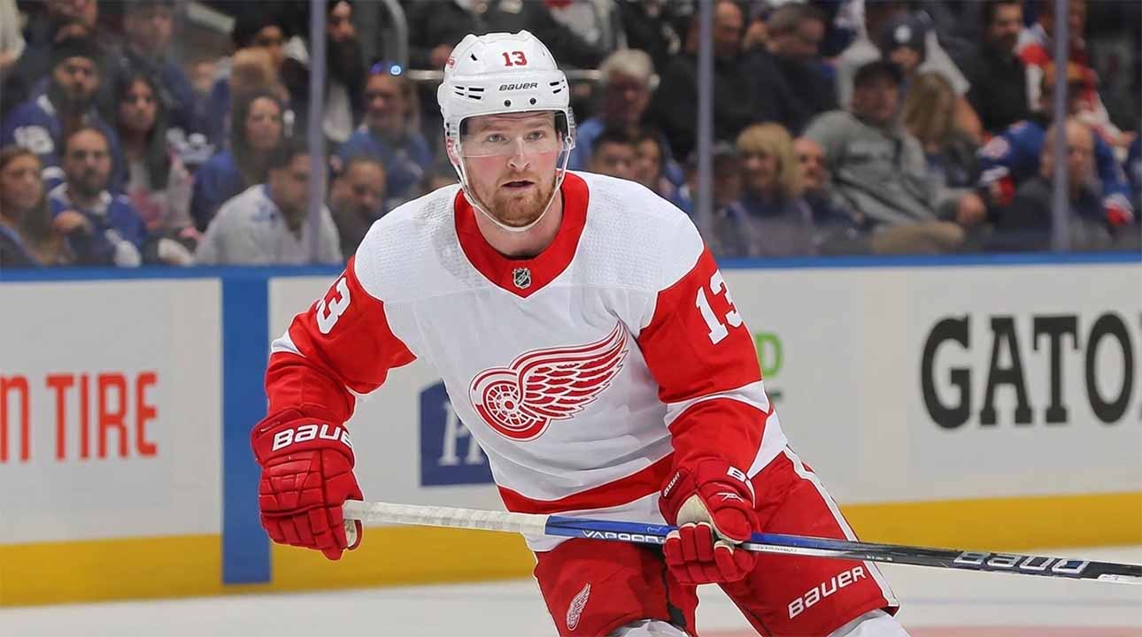 Detroit Red Wings forward Alexis Lafrenière, wearing number 13 in a white away jersey with red trim and the winged wheel logo, skates on the ice holding his stick during a game.
