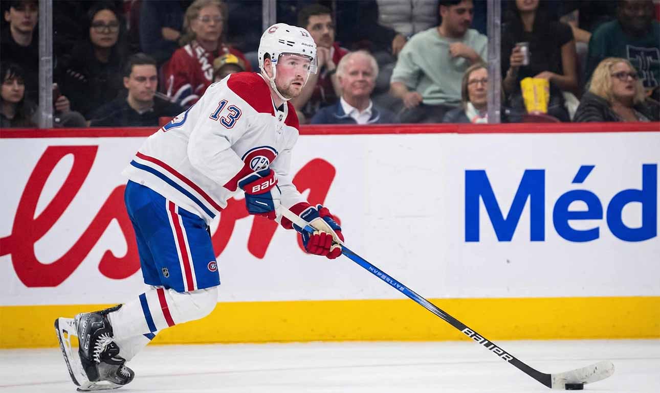 Alexis Lafrenière, wearing a white Montreal Canadiens away jersey with the number 13, skates with the puck on his stick during an ice hockey game. Fans and rink boards with advertisements are visible in the background.