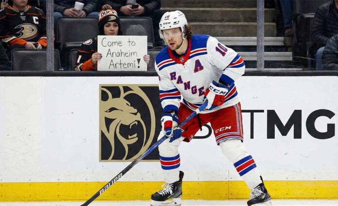 Artemi Panarin, wearing a white New York Rangers away jersey with a blue 'A', is on the ice with his stick during a game. In the background, a fan holds a sign that reads, "Come to Anaheim Artemi!".