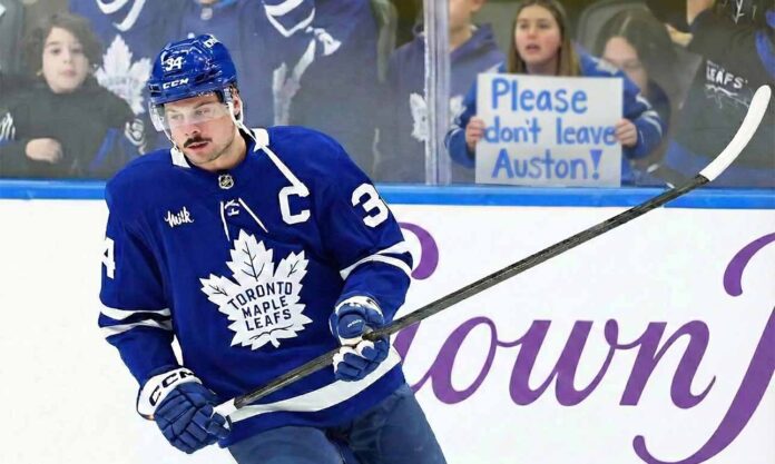 Toronto Maple Leafs captain Auston Matthews skates on the ice during a game, wearing his blue home jersey. Behind the rink glass, a female fan holds up a handwritten white poster board sign with blue text that reads, "Please don't leave Auston!".