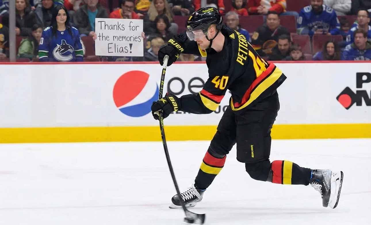 Elias Pettersson (#40) of the Vancouver Canucks, wearing his black "Flying V" jersey, winds up for a slapshot on the ice. Behind him, a female fan with dark hair stands in the front row behind the glass, holding a white sign with handwritten black text that reads, "Thanks for the memories Elias!". Other fans fill the red seats in the background.