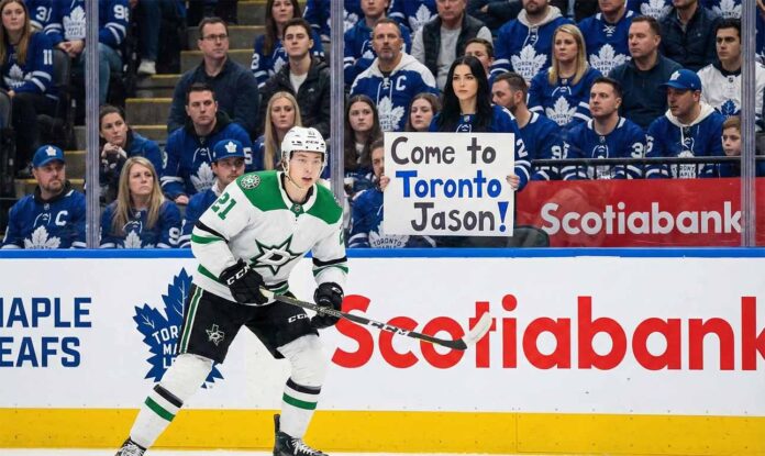 Jason Robertson of the Dallas Stars in his white away uniform skates on the ice during an NHL game against the Toronto Maple Leafs, with fans visible in the stands behind the glass.