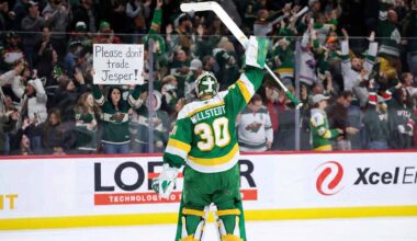 Minnesota Wild goaltender Jesper Wallstedt (30) stands on the ice at Xcel Energy Center, raising his stick to the crowd. Behind the glass boards, a fan holds a handmade sign in red and green ink that reads "Please don't trade Jesper!" amidst cheering spectators.