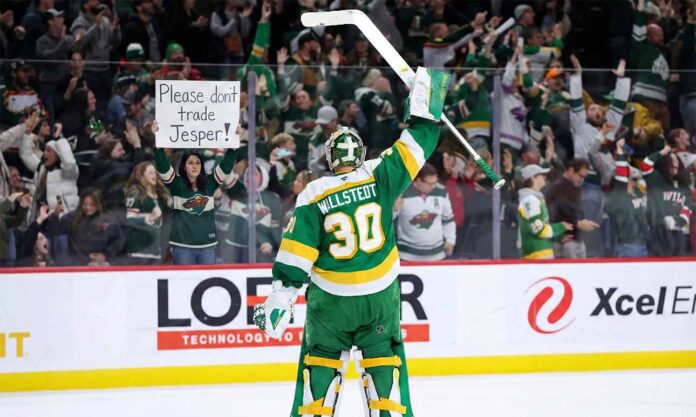 Minnesota Wild goaltender Jesper Wallstedt (30) stands on the ice at Xcel Energy Center, raising his stick to the crowd. Behind the glass boards, a fan holds a handmade sign in red and green ink that reads "Please don't trade Jesper!" amidst cheering spectators.