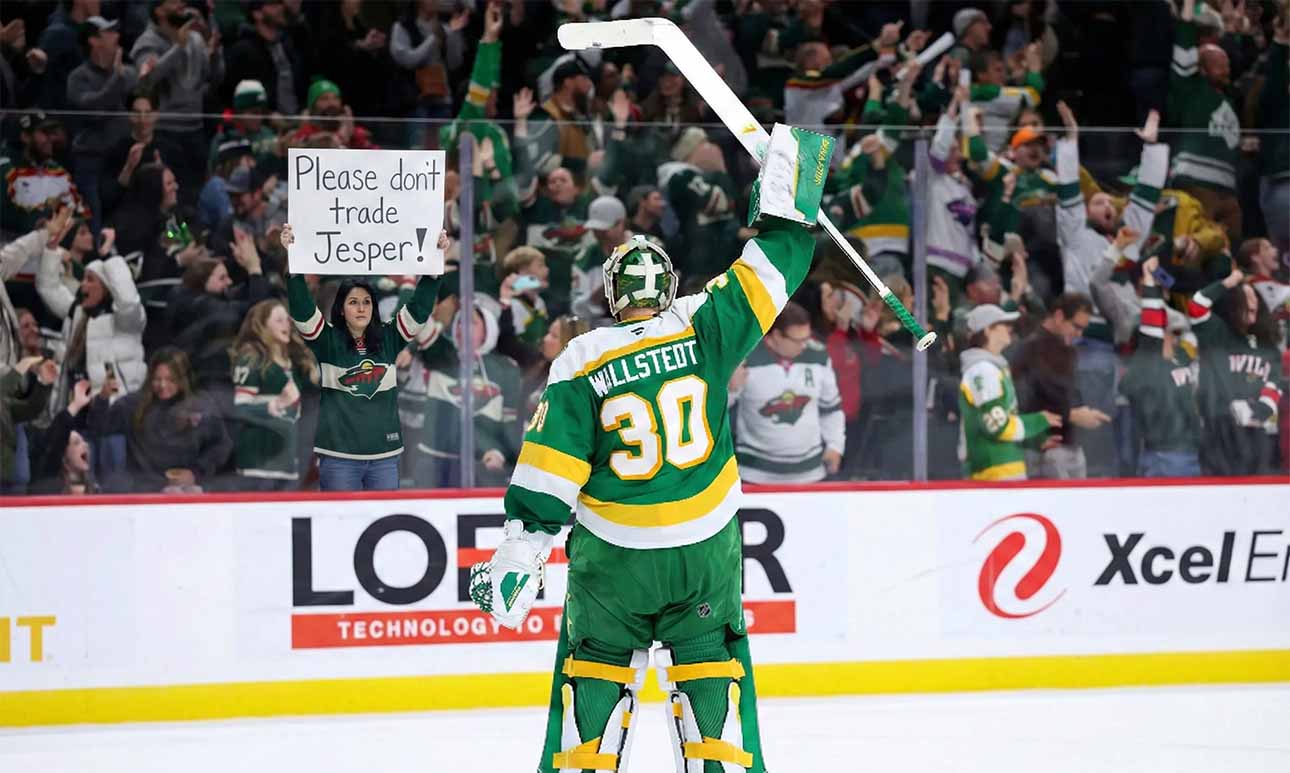 Minnesota Wild goaltender Jesper Wallstedt (30) stands on the ice at Xcel Energy Center, raising his stick to the crowd. Behind the glass boards, a fan holds a handmade sign in red and green ink that reads "Please don't trade Jesper!" amidst cheering spectators.