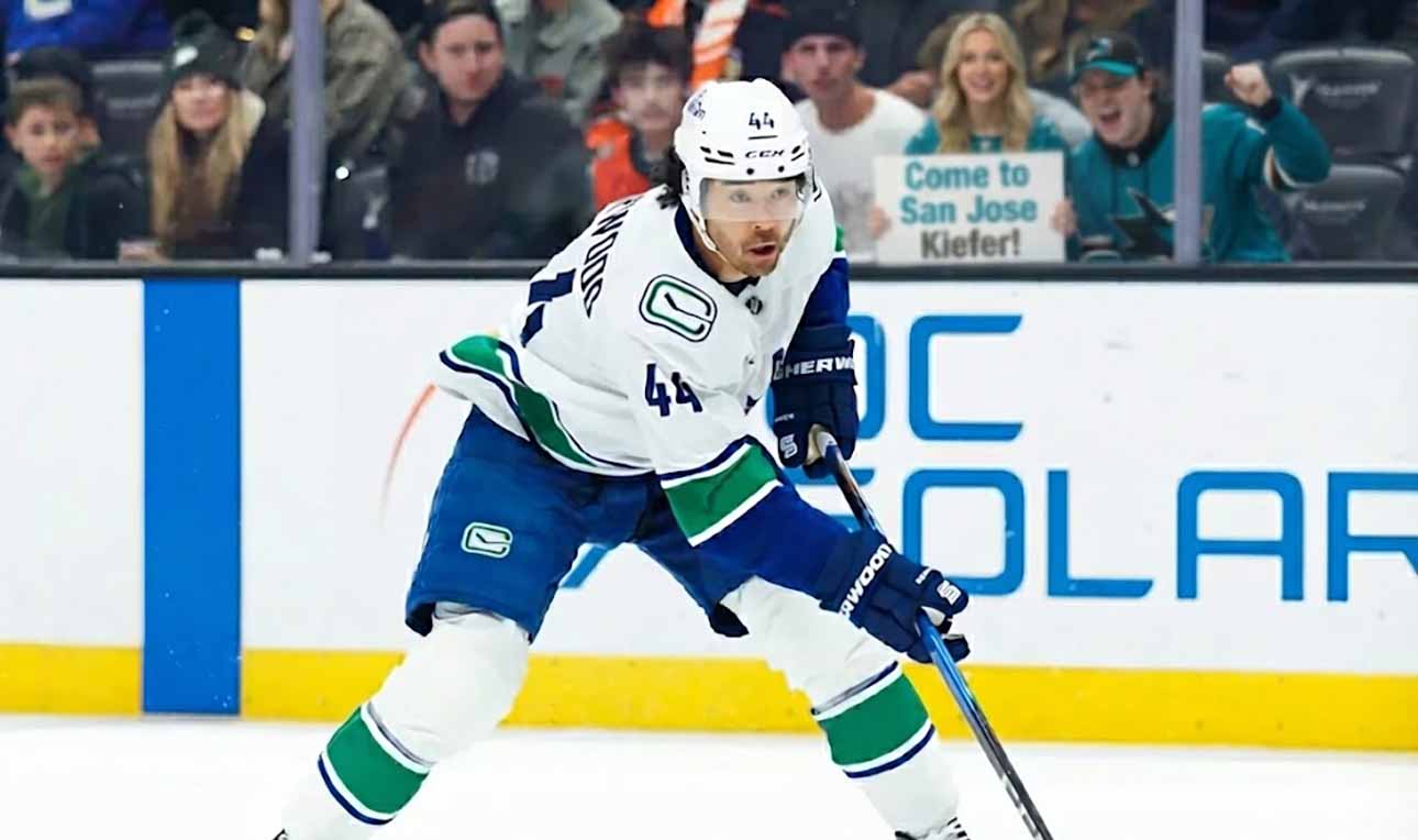 Vancouver Canucks winger Kiefer Sherwood #44 skates with the puck during an NHL game, while San Jose Sharks fans behind the plexiglass hold a sign reading "Come to San Jose Kiefer!".
