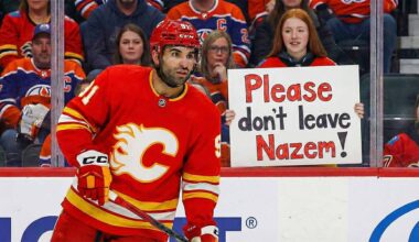 Calgary Flames center Nazem Kadri, wearing his red home jersey, looks towards the action on the ice. In the stands behind the glass, a young female fan with red hair holds up a handwritten sign with purple and orange letters that reads, "Please don't leave Nazem!".