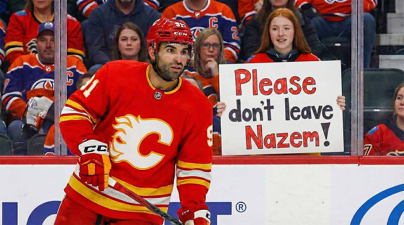 Calgary Flames center Nazem Kadri, wearing his red home jersey, looks towards the action on the ice. In the stands behind the glass, a young female fan with red hair holds up a handwritten sign with purple and orange letters that reads, "Please don't leave Nazem!".