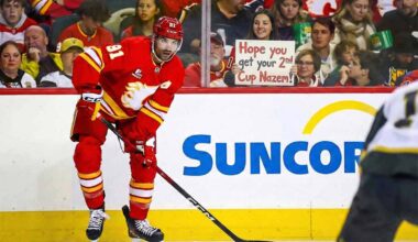 Calgary Flames center Nazem Kadri (#91) skating on the ice during a game, looking towards the camera. Behind the glass in the background, a smiling fan holds up a handwritten sign that reads, "Hope you get your 2nd Cup Nazem!"