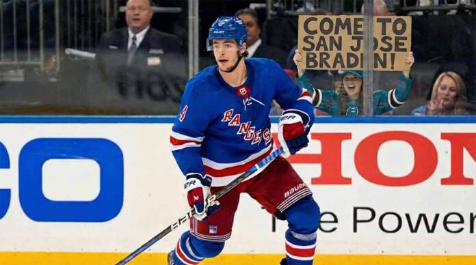 New York Rangers defenseman Braden Schneider (#4) skates on the ice during a game, while a female fan wearing San Jose Sharks apparel behind the glass holds up a handmade cardboard sign that reads, "COME TO SAN JOSE BRADEN!".