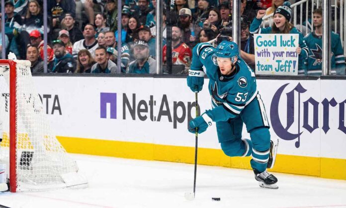 San Jose Sharks forward Jeff Skinner (#53) skates with the puck during a game. Behind him in the stands, a smiling female fan wearing a Sharks beanie holds a handwritten sign that reads, 'Good luck with your next team Jeff!'.