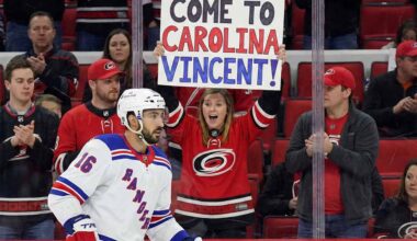 Vincent Trocheck, wearing a New York Rangers jersey, skates on the ice during a game, while a female fan in a Carolina Hurricanes jersey behind the glass holds a sign that reads, "COME TO CAROLINA VINCENT!".