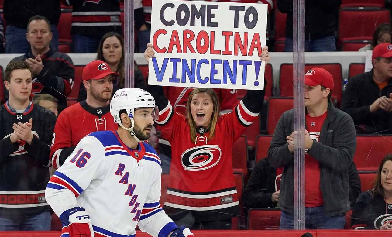 Vincent Trocheck, wearing a New York Rangers jersey, skates on the ice during a game, while a female fan in a Carolina Hurricanes jersey behind the glass holds a sign that reads, "COME TO CAROLINA VINCENT!".