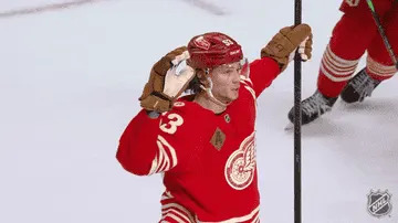 Hockey player celebrates a goal on the ice, raising a hand to his ear, looking toward the audience, holding a hockey stick in the other hand
