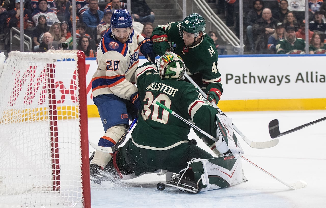 A scrum of men's hockey players happens in front of a goalie, in a forest green uniform. The puck is loose beneath his pads.
