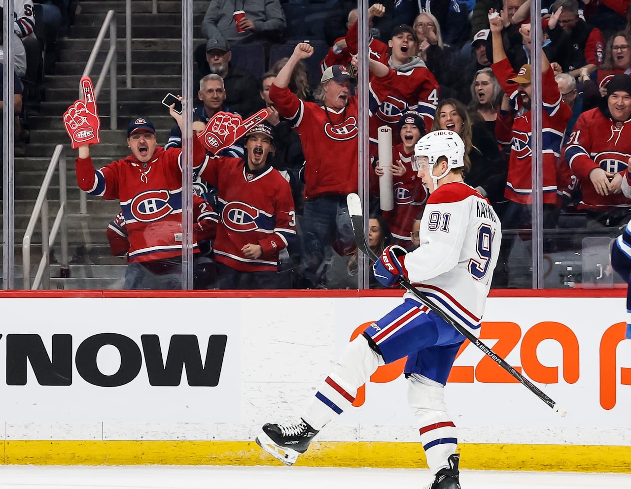 A hockey player in a white jersey with red and blue trim celebrates a goal. Fans behind the glass cheer.