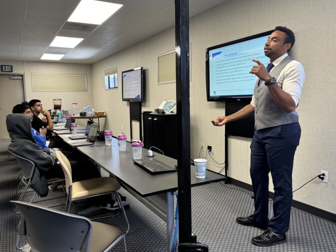 A middle aged man with dark skin and short black hair dressed in a suit speaks to a handful of students in a room. 