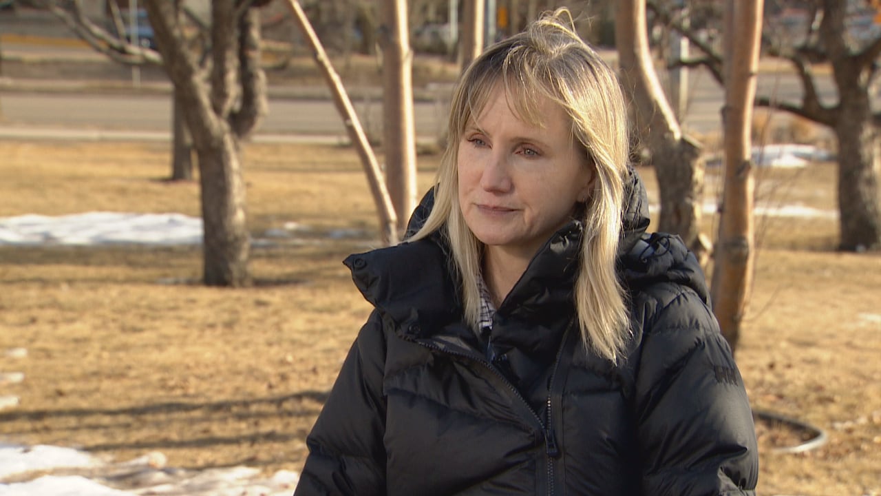A woman with blonde hair wears a black jacket. She is outside. The grass is dead and there is snow on the ground. She has tears in her eyes.