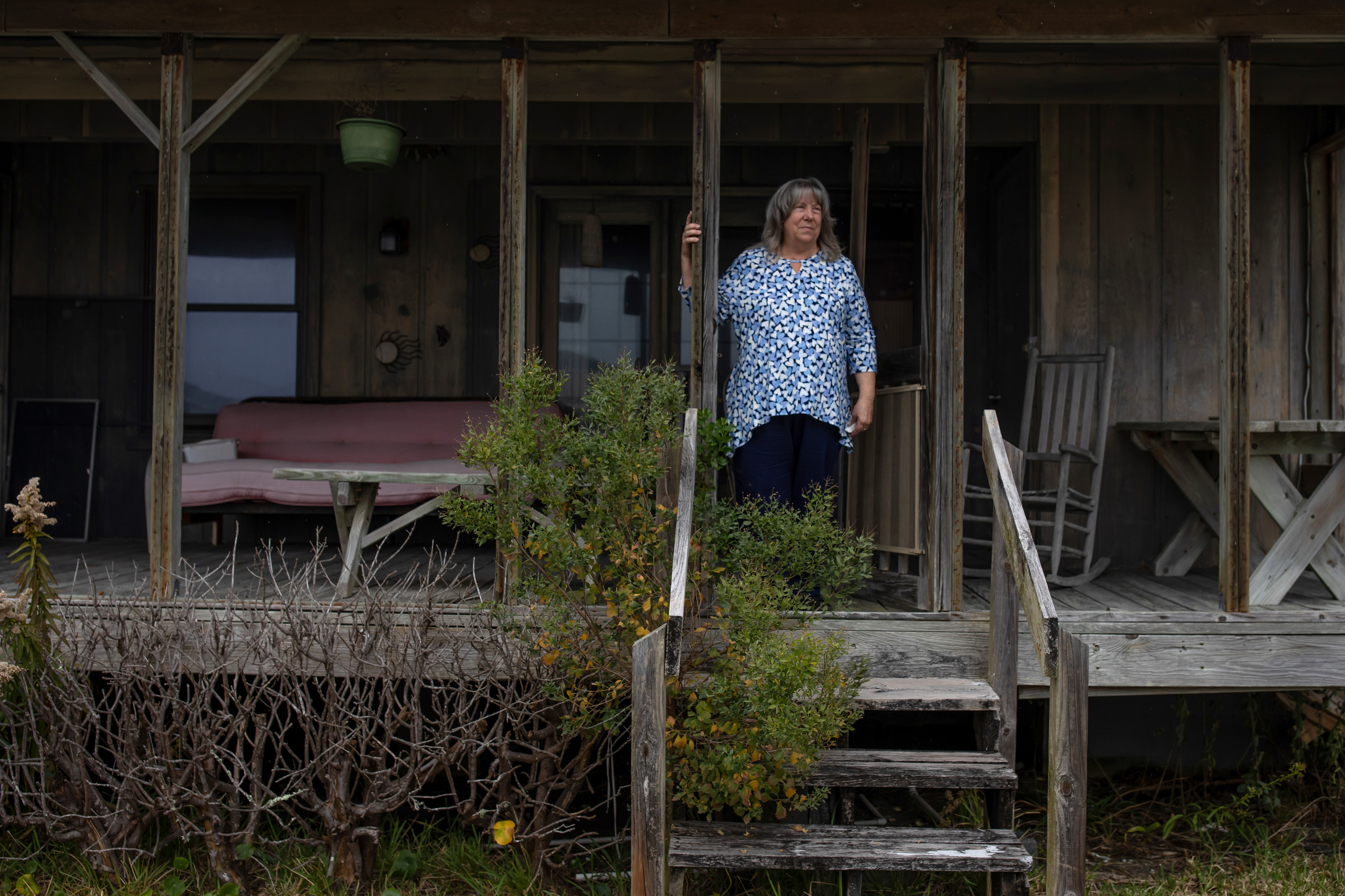 A woman wearing a blue floral shirt stands on the porch of a weathered wooden house.