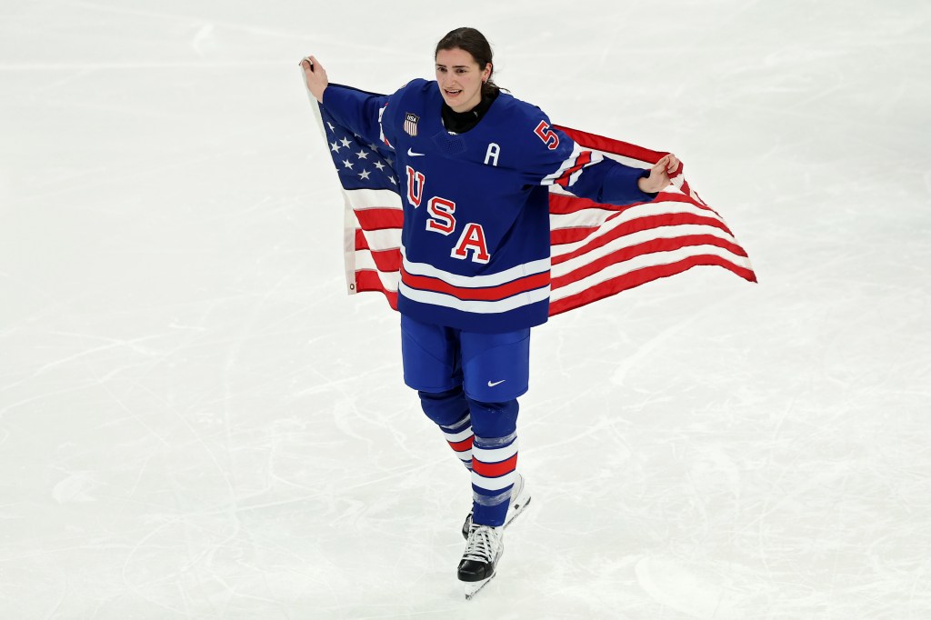 US women's ice hockey player in a blue jersey with "USA" and an "A" on it, skating on ice while holding an American flag behind her back.