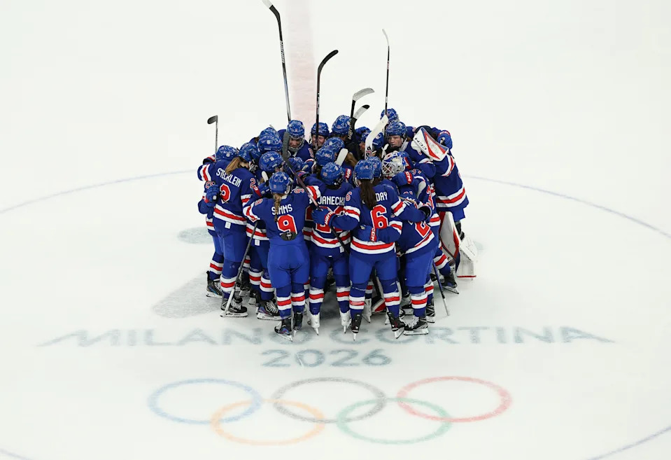 Team in blue hockey uniforms huddles on ice above "Milano Cortina 2026" and Olympic rings