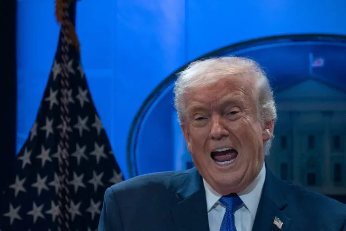 Person in a suit speaking energetically on stage with a flag and emblem visible in the background