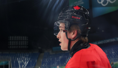 A hockey player in a red jersey and black helmet with a red maple leaf watches intently on an Olympic ice rink.