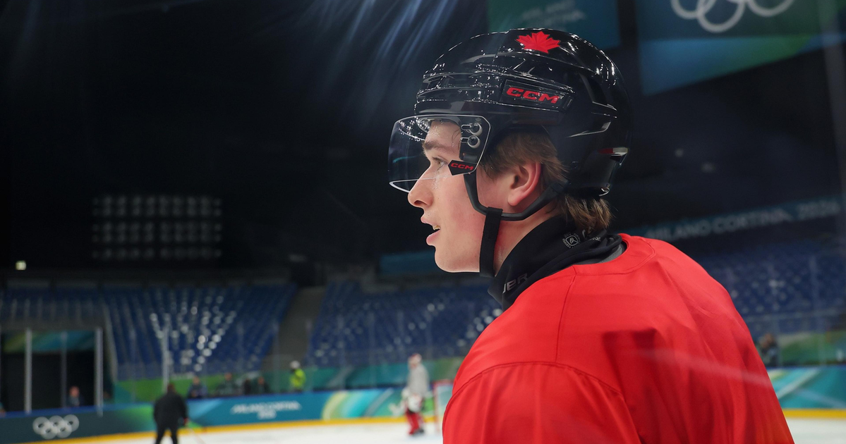 A hockey player in a red jersey and black helmet with a red maple leaf watches intently on an Olympic ice rink.