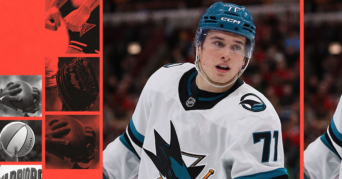 A hockey player wearing a white San Jose Sharks jersey with number 71 and a blue helmet skates on the ice with a focused expression.