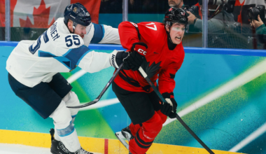 A hockey player in a red uniform skates quickly while controlling the puck, chased closely and grabbed from behind by an opponent in a white and blue uniform.
