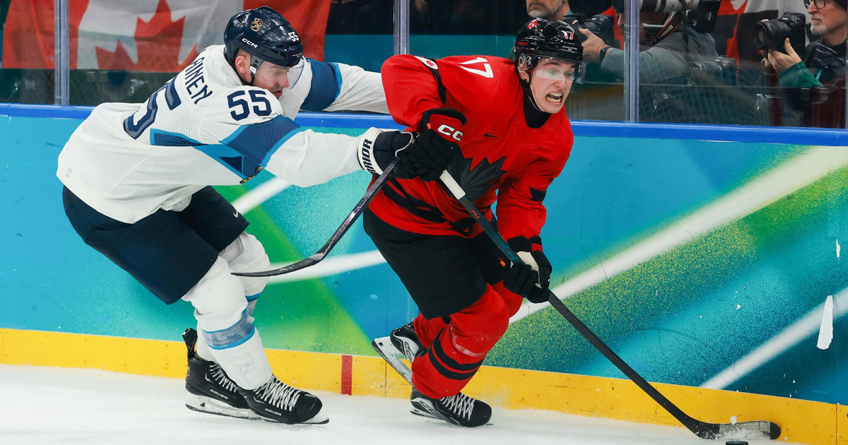 A hockey player in a red uniform skates quickly while controlling the puck, chased closely and grabbed from behind by an opponent in a white and blue uniform.
