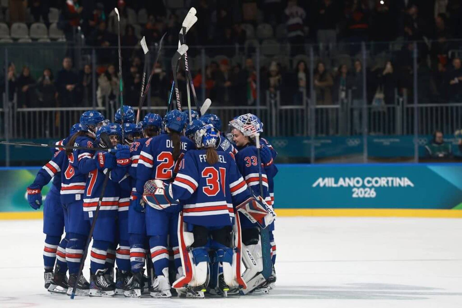 The United States women's hockey team, wearing blue uniforms, huddling together and raising their sticks in the air.