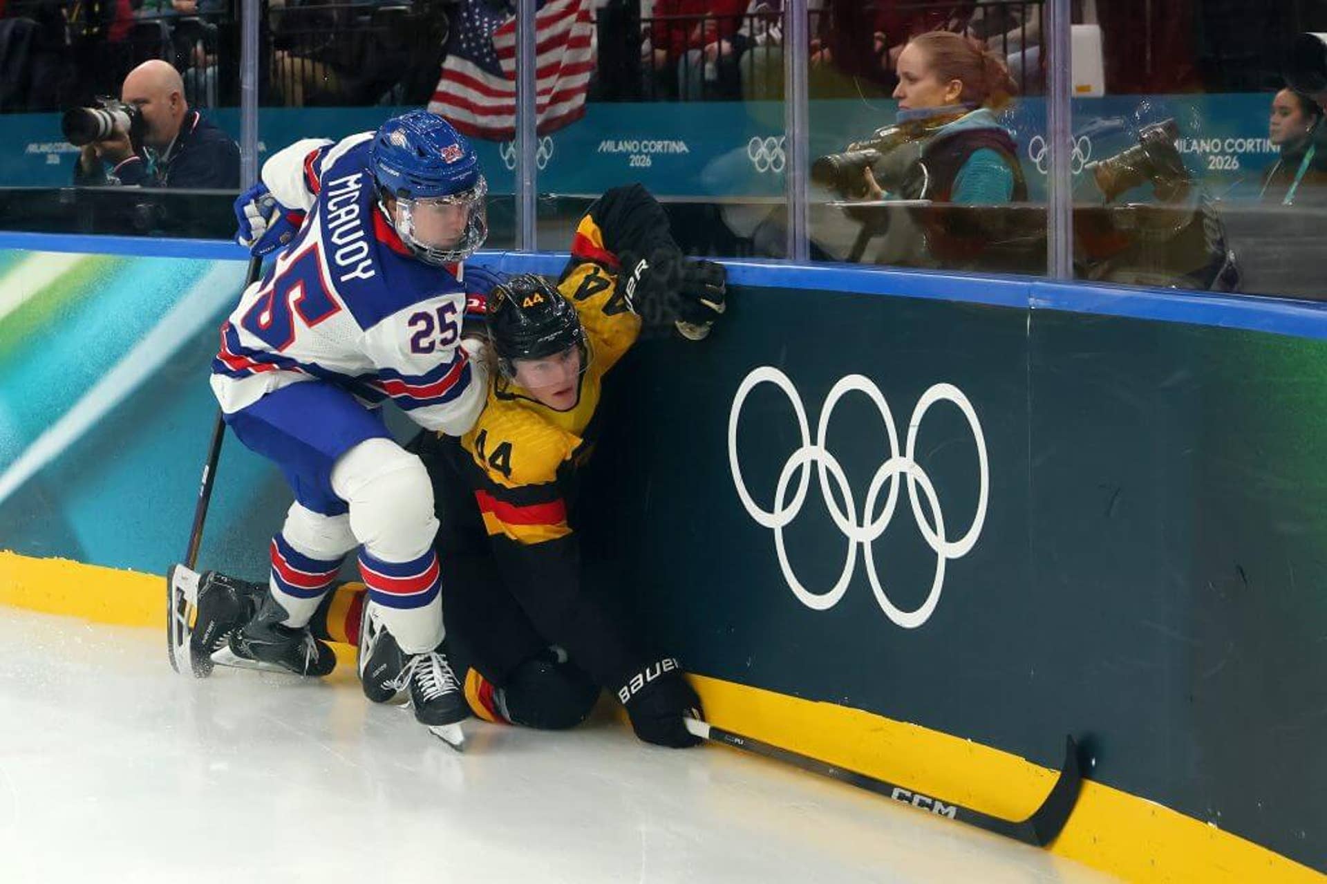 Team USA's Charlie McAvoy and Germany's Josh Samanski collide near the boards, which are a dark turquoise green.