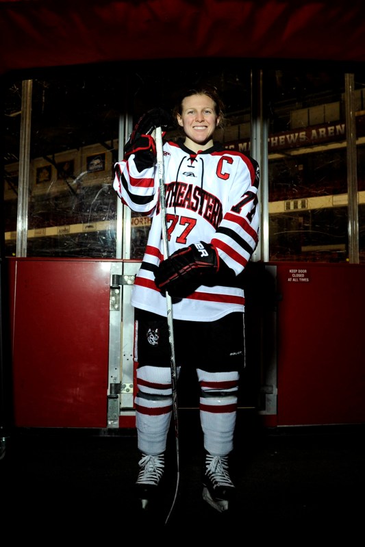 Kendall Coyne Schofield smiles while standing on the ice in full Northeastern garb.
