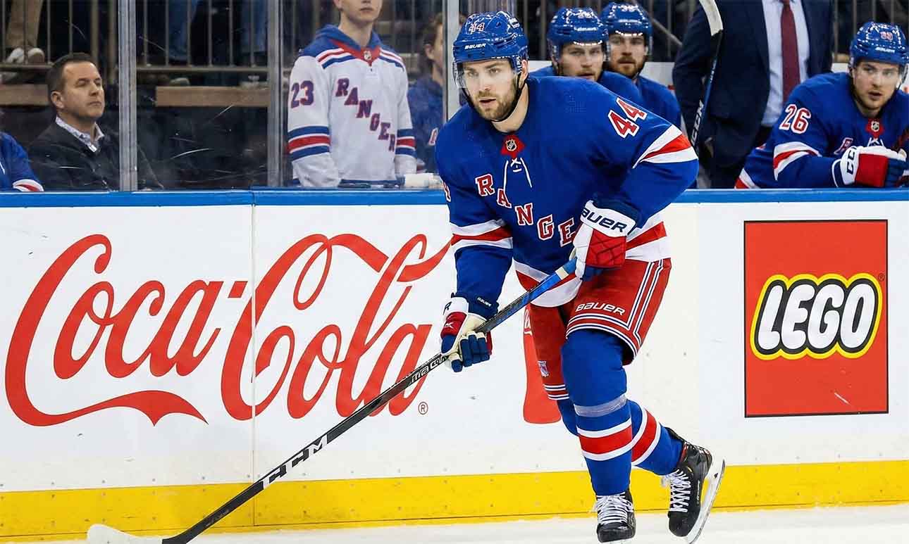 NY Rangers defenseman Vladislav Gavrikov (#44), currently the subject of intense trade rumors amid reports he is unhappy with the team's rebuild, skates with the puck past Coca-Cola and LEGO advertisements on the boards.