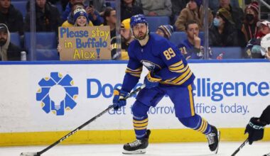 Buffalo Sabres forward Alex Tuch, wearing number 89 in the blue home jersey, skates with the puck during a game. Behind him, in the spectator stands, a female fan wearing a Sabres winter hat holds up a cardboard sign with blue and yellow lettering that reads, "Please stay in Buffalo Alex!".