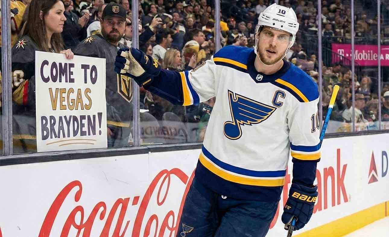 St. Louis Blues captain Brayden Schenn, wearing his white away jersey with the 'C' patch, skates along the boards at T-Mobile Arena, looking towards a female fan behind the glass holding a handmade sign that reads "COME TO VEGAS BRAYDEN!".