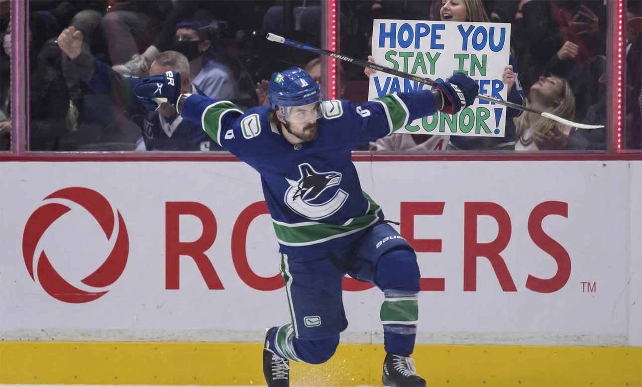 Vancouver Canucks forward Conor Garland (#8) celebrates a goal by sliding on one knee across the ice with his arms extended. Behind the glass boards featuring the Rogers logo, cheering fans are visible, including a woman holding up a handmade white sign with blue and green lettering that reads, "HOPE YOU STAY IN VANCOUVER CONOR!".