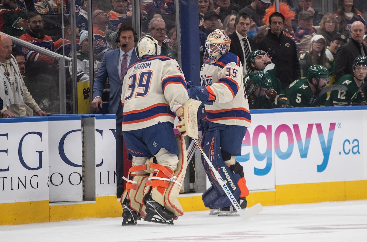 Two men's hockey goalies, in full white-blue-and-orange uniform, sub out for each other near the bench.