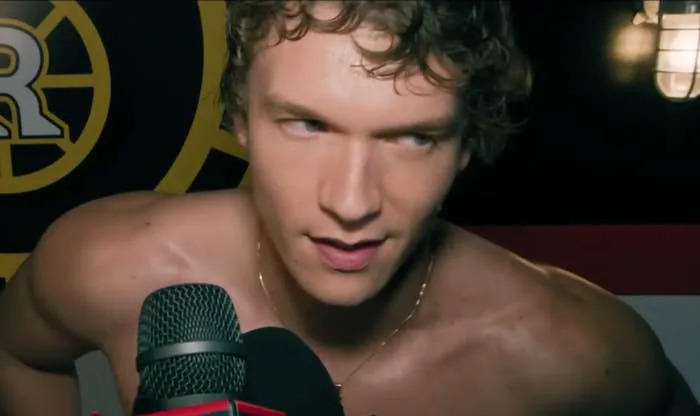 Connor Storrie speaks into microphones, looking sideways, with a sports team logo partially visible in the background as Ilya on Heated Rivalry