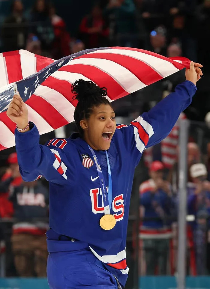 Athlete celebrates joyfully with a medal and a large national flag draped over shoulders, wearing a sports uniform with "USA" lettering