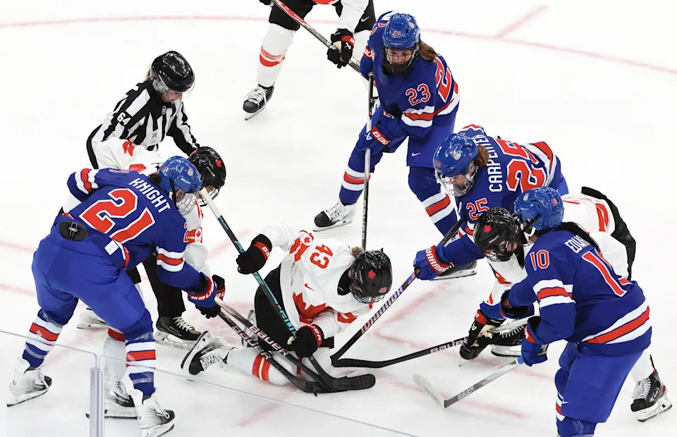 Hockey players from two teams, in red and white uniforms, compete for the puck on the ice, surrounded by a referee watching closely