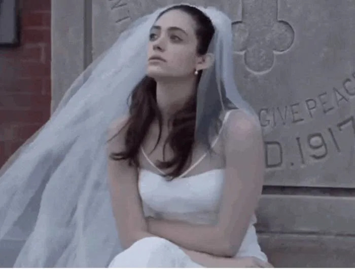 A person in a wedding dress with a veil sits pensively in front of a gravestone