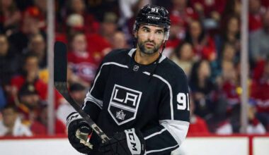 Nazem Kadri depicted wearing a black Los Angeles Kings number 91 hockey jersey, holding his stick and looking towards the right side of the frame against a blurred arena crowd background.