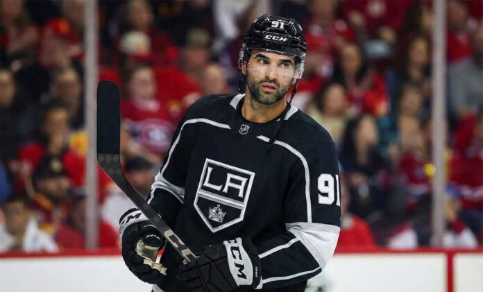 Nazem Kadri depicted wearing a black Los Angeles Kings number 91 hockey jersey, holding his stick and looking towards the right side of the frame against a blurred arena crowd background.