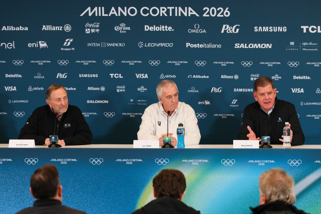 Three men, identified as Gary Bettman, Luc Tardif, and Marty Walsh, sit at a table before a backdrop displaying various sponsor logos for the Milano Cortina 2026 Winter Olympics.