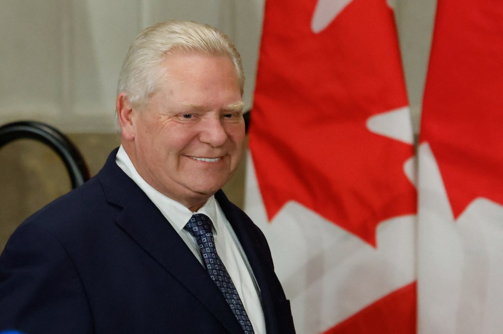 Ontario Premier Doug Ford smiles, wearing a dark suit and patterned tie, with Canadian flags in the background.