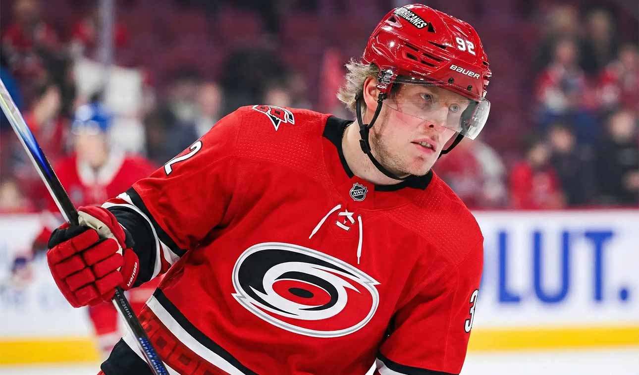 A close-up photograph of professional ice hockey player Patrik Laine wearing a red Carolina Hurricanes jersey with the team's logo, a red helmet with "HURRICANES" and number 92, and red gloves. He is holding a hockey stick and