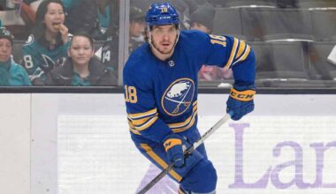 Robert Thomas wearing a blue Buffalo Sabres jersey with the number 18, blue helmet, blue gloves, and holding a hockey stick, is skating on the ice during a game. Spectators are visible in the background behind the glass boards.